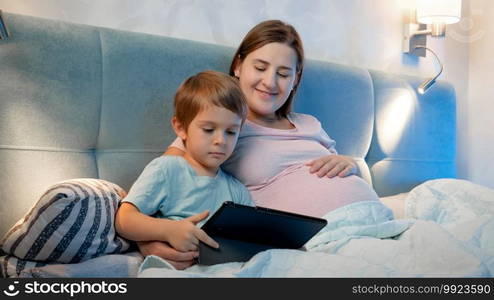 Family lying in bed and watching cartoons on tablet computer at night. Little boy and pregnant mother using digital tablet in bed at night.. Family lying in bed and watching cartoons on tablet computer at night. Little boy and pregnant mother using digital tablet in bed at night