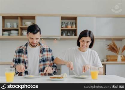 Family married couple pose at kitchen table, have delicious breakfast, talk about plannings on day, eat fried eggs and burgers, drink fresh apple juice, dressed casually, enjoy domestic atmosphere