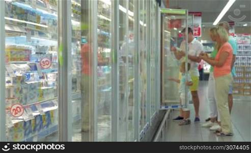 Family members are standing by the refrigerator in the store, opening the doors and choosing goods. Shot is slightly defocused for adding your content on the foreground.