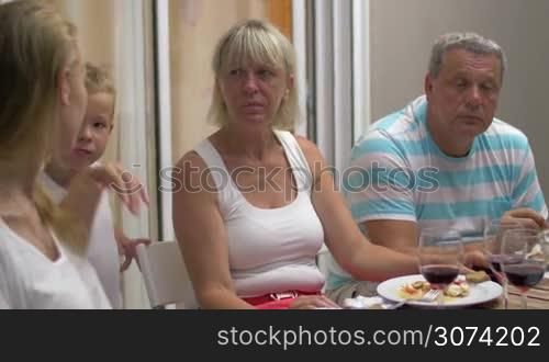 Family members including senior people and young woman talking vividly during dinner. Child coming up to kiss dear mother
