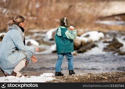 Family - mother and son to be seen - on a walk along a riverbank in winter; the child is throwing a snowball