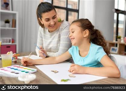 family, motherhood and leisure concept - happy smiling mother spending time with her little daughter drawing or painting wooden chipboard items with colors at home. happy mother with little daughter drawing at home