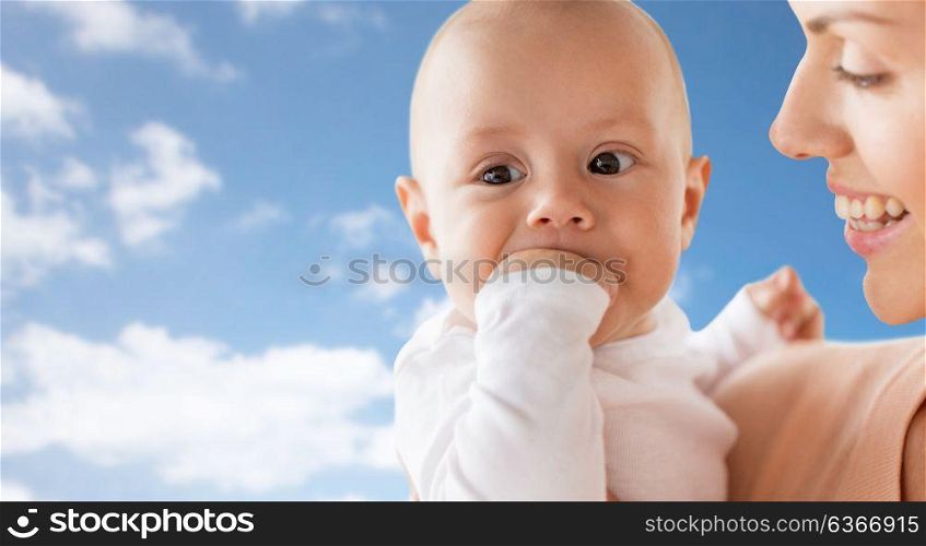 family, motherhood and people concept - close up of happy mother with little baby boy sucking fingers over blue sky and clouds background. happy mother with baby sucking fingers over sky