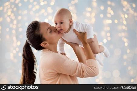 family, motherhood and people concept - happy mother kissing little baby boy over holidays lights background. happy mother kissing little baby boy over lights