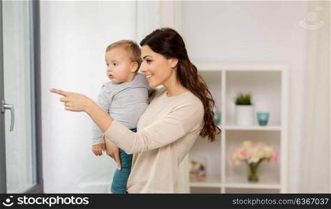 family, motherhood and people concept - happy mother with little baby boy looking through window at home. happy mother with little baby boy at home