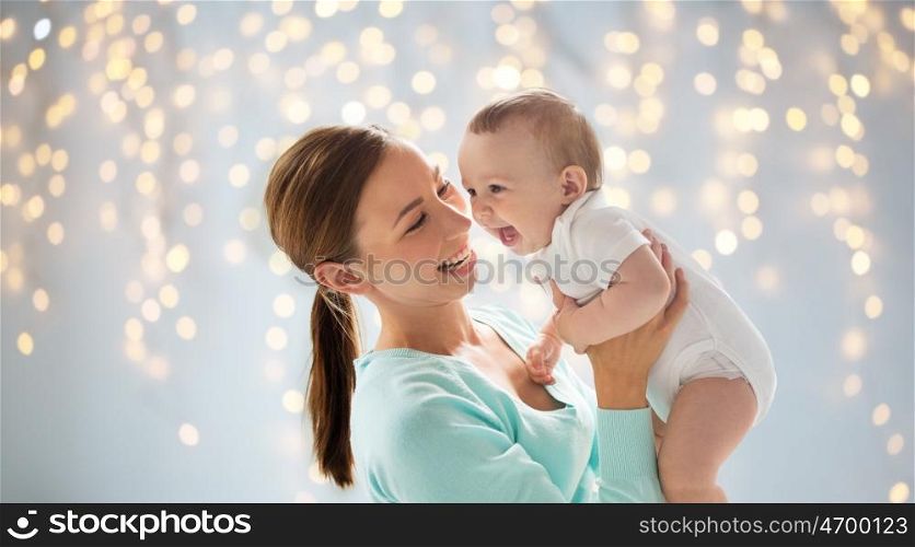 family, motherhood, child and parenthood concept - happy smiling young mother with little baby over holidays lights background