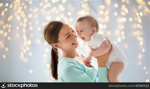 family, motherhood, child and parenthood concept - happy smiling young mother with little baby over holidays lights background