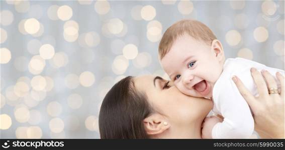 family, motherhood, children, parenthood and people concept - happy mother kissing her baby over holidays lights background
