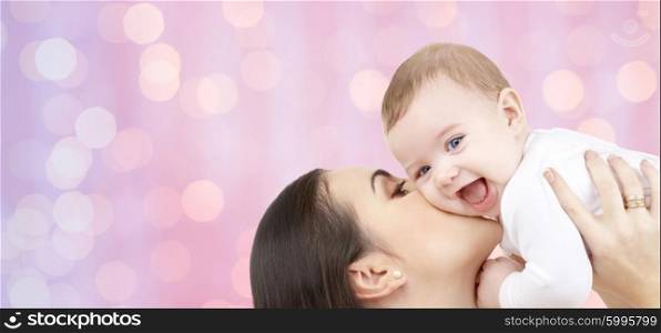 family, motherhood, children, parenthood and people concept - happy mother kissing her baby over pink holidays lights background