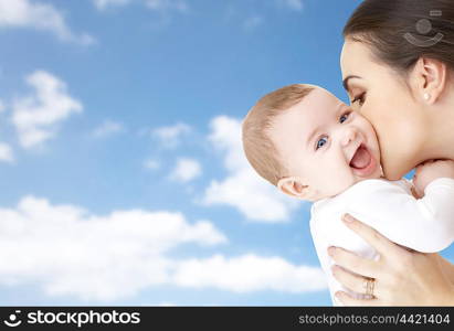 family, motherhood, parenting, people and child care concept - happy mother kissing adorable baby over blue sky background