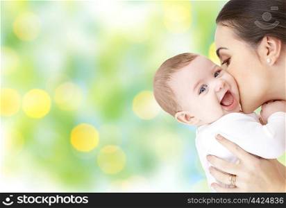 family, motherhood, parenting, people and child care concept - happy mother kissing adorable baby over green holidays lights background