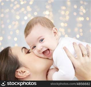 family, motherhood, parenting, people and child care concept - happy mother kissing adorable baby over holidays lights background