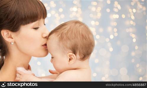 family, motherhood, parenting, people and child care concept - happy mother kissing adorable baby over holidays lights background