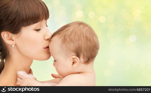 family, motherhood, parenting, people and child care concept - happy mother kissing adorable baby over green holidays lights background