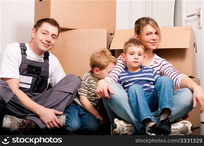 Family moving in their new house. They are sitting in front of a stack of moving boxes being happy. Father is dressed in a way that can also represent a mover