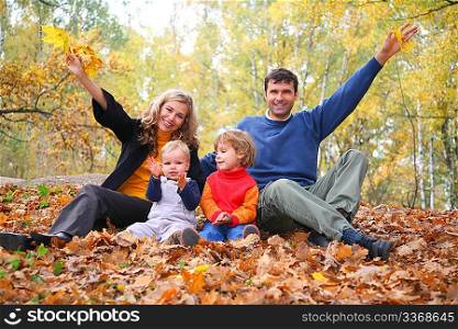 family of four sits in autumn park. parent hands up.