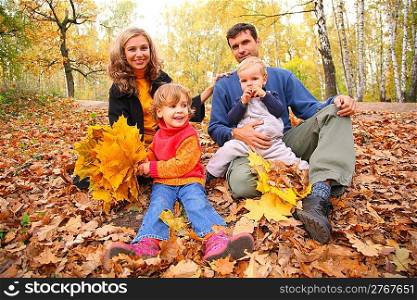 Family of four with yellow maple leaves sits in wood in autumn