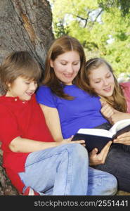 Family of mother and children reading a book under a tree in summer park