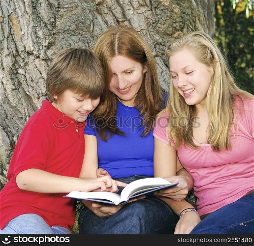 Family of mother and children reading a book under a tree in summer park
