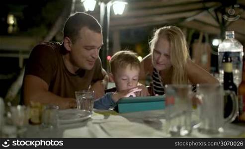 Family of three in outdoor cafe in the evening. Boy playing on touch pad, happy parents watching