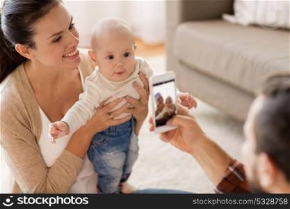 family, parenthood and people concept - happy father with smartphone taking picture of mother with baby at home. happy family with baby photographing at home