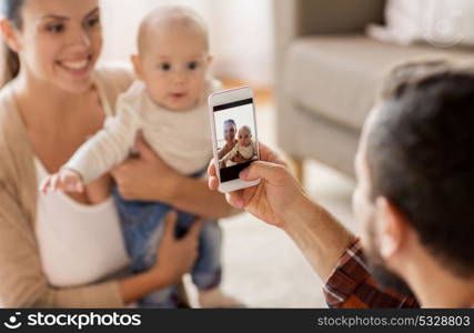 family, parenthood and people concept - happy father with smartphone taking picture of mother with baby at home. happy family with baby photographing at home