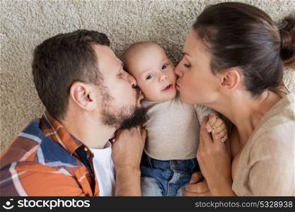 family, parenthood and people concept - happy mother and father lying on floor and kissing their baby at home. happy family lying on floor and kissing their baby