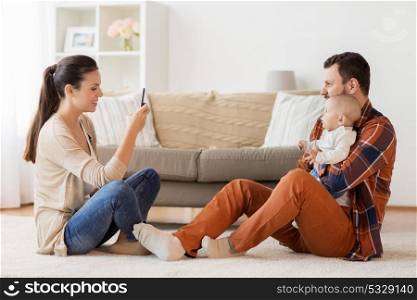 family, parenthood and people concept - happy mother with smartphone taking picture of father with baby at home. happy family with baby photographing at home