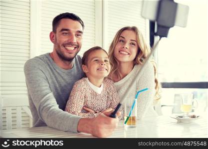 family, parenthood, technology and people concept - happy mother, father and little girl having dinner and taking picture by smartphone selfie stick at restaurant