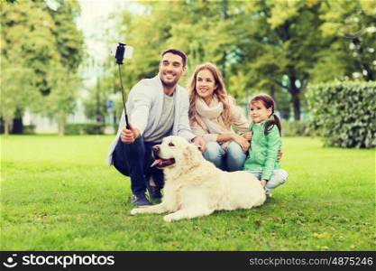 family, pet, animal, technology and people concept - happy family with labrador retriever dog taking picture by smartphone on selfie stick in park. happy family with dog taking selfie by smartphone