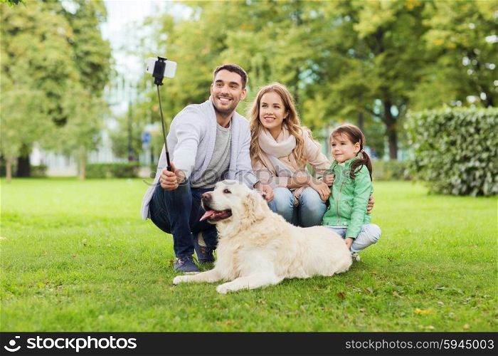 family, pet, animal, technology and people concept - happy family with labrador retriever dog taking picture by smartphone on selfie stick in park