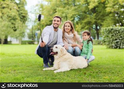 family, pet, animal, technology and people concept - happy family with labrador retriever dog taking picture by smartphone on selfie stick in park