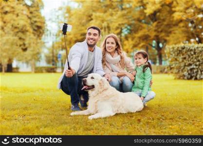 family, pet, season, technology and people concept - happy family with labrador retriever dog taking picture by smartphone on selfie stick in autumnl park