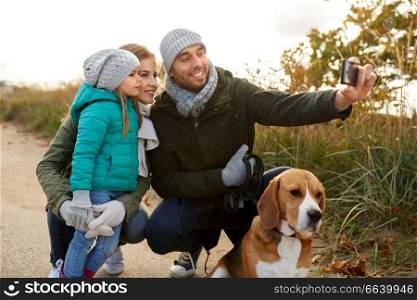 family, pets and people concept - happy mother, father and little daughter with beagle dog taking selfie by smartphone outdoors in autumn. happy family with dog taking selfie in autumn