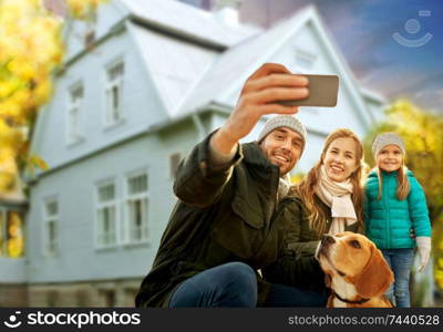 family, pets and people concept - happy mother, father and little daughter with beagle dog taking selfie by smartphone outdoors over house in autumn background. family with dog taking selfie over house in autumn