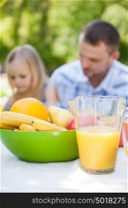 Family picnic. Father and daughter sitting together and talking on background. Fruits and juice on foreground. Focus on food