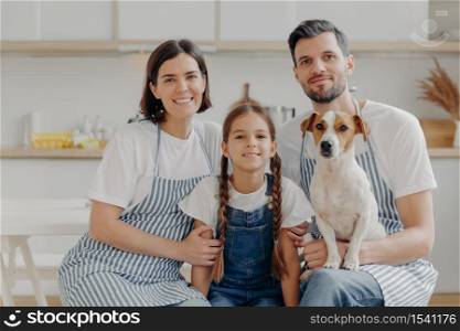 Family portrait of father, mother, daughter and pedigree dog pose together for making memorable photo, have rest after preparing dinner, poses against kitchen interior, have happy expressions