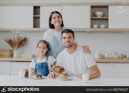 Family portrait of happy mother, daughter and father pose at kitchen during breakfast time, eat delicious homemade pancakes, their dog poses near, have friendly good relationships, love each other