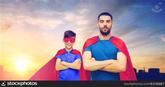 family, power and people concept - father and son in red superhero capes over city and sun light background. father and son in superhero capes over city