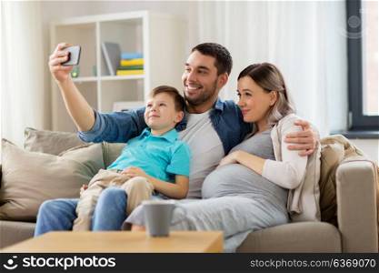 family, pregnancy and technology concept - happy pregnant mother, father and little son taking selfie with smartphone sitting on on sofa at home. happy family taking selfie with smartphone at home