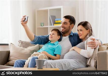 family, pregnancy and technology concept - happy pregnant mother, father and little son taking selfie with smartphone sitting on on sofa at home. happy family taking selfie with smartphone at home. happy family taking selfie with smartphone at home