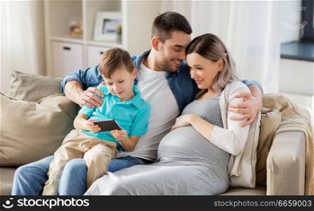 family, pregnancy and technology concept - happy pregnant mother, father and little son with smartphone sitting on on sofa at home. happy family with smartphone at home