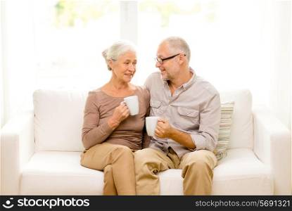 family, relations, age, drinks and people concept - happy senior couple with cups hugging on sofa at home