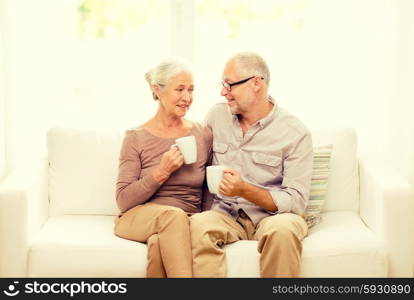 family, relations, age, drinks and people concept - happy senior couple with cups hugging on sofa at home