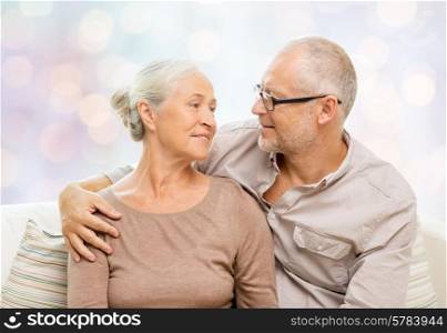 family, relations, love, age and people concept - happy senior couple hugging and looking at each other on sofa over holidays lights background