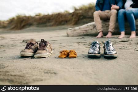 Family shoes in the sand with pregnant woman in the background with her partner. Family shoes in the sand