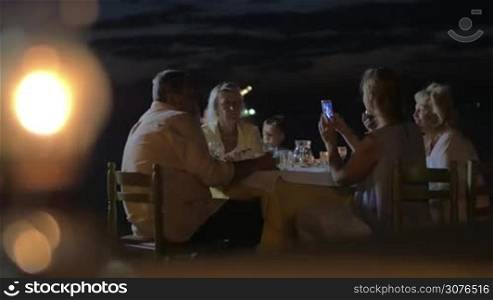 Family sitting at a table in evening near the coast and having dinner in city Perea, Greece. Girl is photographed on a mobile phone her mother and father. Happy family resting together