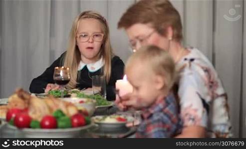 Family sitting at festive table served with tasty roasted turkey and delicious dishes on Thanksgiving holiday. Lovely little girl eating and telling story to her little brother. Cute toddler boy sitting on grandmother knees.