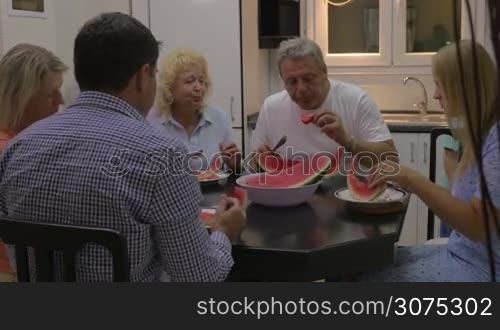 Family sitting at the dining table and eating sweet watermelon, child walking around and mother feeding him