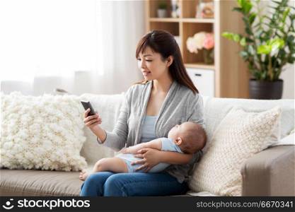 family, technology and motherhood concept - happy smiling young asian mother with sleeping baby and smartphone at home. mother with sleeping baby and smartphone at home. mother with sleeping baby and smartphone at home